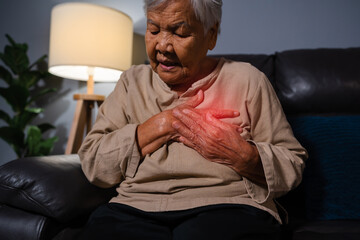 senior woman suffering from bad pain in her chest heart while sitting on sofa in the living room at night