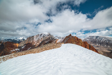 Snowfield on stony mountain ridge and vivid sharp pinnacle in sunlight. Shiny pointy peak of gold color. Colorful red rocky peaked top. Freshly fallen snow in high mountains. Sunny cloudy alpine view. - 794639824