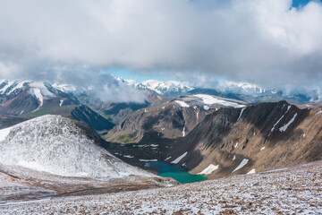 Scenic view from above to most beautiful turquoise alpine lake among hills in freshly fallen snow and large snowy mountains in low clouds. High mountain range with snow-white peaks in low cloudy sky.