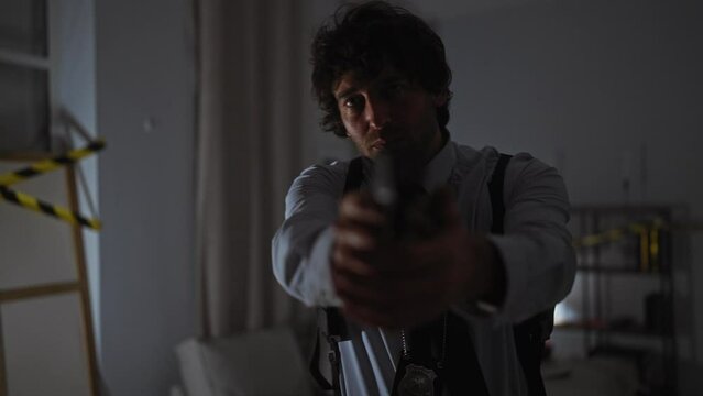 An intense young man in a shirt and tie with a detective badge investigates a dim, indoor crime scene, pointing a gun.