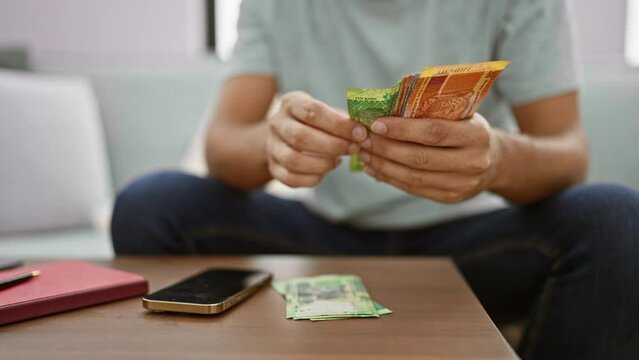 Young hispanic man chilling on sofa, happily counting his stash of south african rand banknotes, busy texting about his flourishing business from his cosy living room
