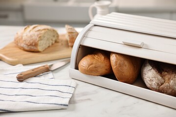 Wooden bread basket with freshly baked loaves and knife on white marble table in kitchen
