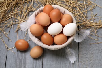 Fresh chicken eggs in bowl, feathers and dried straw on grey wooden table, above view