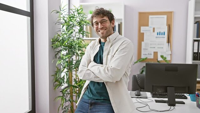A confident man in glasses stands with arms crossed in a modern office with a computer and plants in the background.