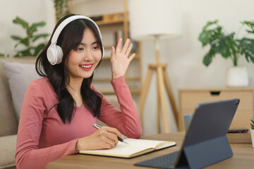 Young businesswoman greeting with partners and writing note in notebook while meeting on video call