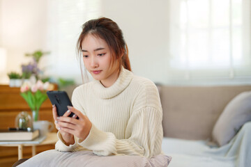 Young asian women in sweater enjoying with surfing entertainment on smartphone while sitting on comfortable couch to relaxation and doing activity for spending time with slow life lifestyle at home