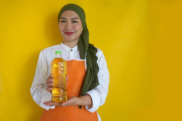 Adult Asian muslim woman holding and showing a cooking oil bottle on yellow background