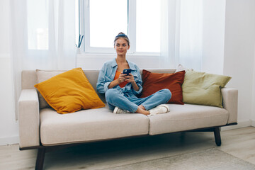 Happy Young Woman Relaxing on the Sofa, Holding a Mobile Phone and Smiling in her Cozy Living Room