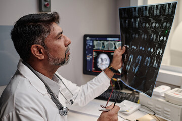 Mature male radiographer sitting at table in office working with head X-ray scan, medium closeup