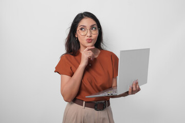 Thoughtful young woman wearing brown shirt and eyeglasses looking aside while holding laptop...