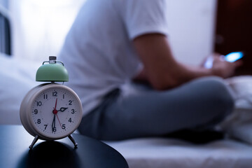 Asian sleepless man individual sit in bed browsing their smartphone with an alarm clock in the...
