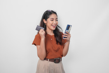 Beautiful young Asian woman wearing brown shirt is smiling while holding her smartphone and credit card over isolated white background.