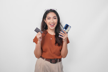 Happy successful Asian woman wearing brown shirt is holding her smartphone and credit card over isolated white background.