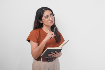 Thoughtful young Asian woman wearing a brown shirt and eyeglasses holding a book and looking aside...
