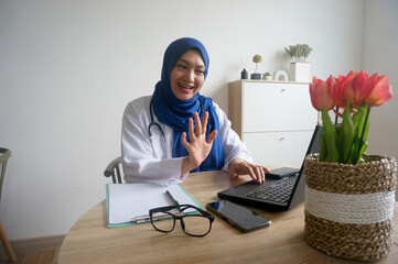Asian female doctor in hijab waving hand say hi and having online consultation on digital tablet...