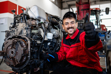 Portrait of mechanic holding thumbs up and servicing tractor engine in workshop.
