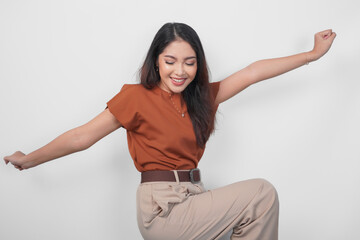 Cheerful Asian woman in brown shirt dancing with excited expression and having fun gesture over isolated white background.