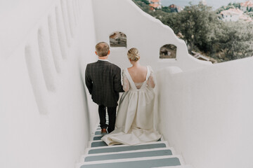 A bride and groom are walking down a white staircase. The bride is wearing a white dress and the...