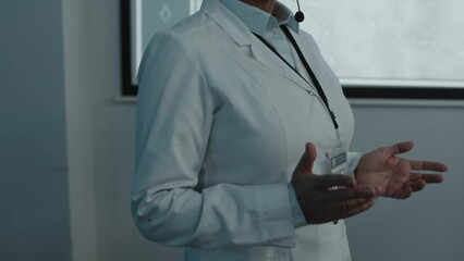 Female doctor wearing white coat and microphone headset walking in front of projection screen and explaining presentation on medical conferenceFemale doctor wearing white coat and microphone headset w
