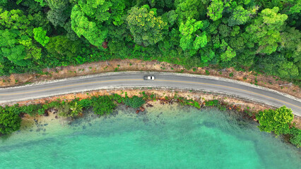 Cars cruise along a breathtaking tropical coastal road, bordered by lush greenery and crystal-clear...