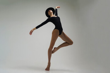 young ballerina in a black bodysuit shows ballet steps in motion standing on one leg and spreading...