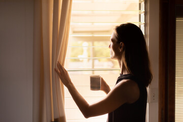Young woman looking out her window early morning enjoying a cup of coffee to start a new day - obrazy, fototapety, plakaty