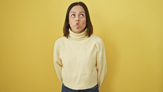 Zany young hispanic woman making a comical fish face and mad gesture while joyfully standing solo, rocking her beautiful sweater against an isolated yellow background
