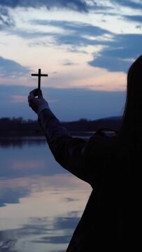 The silhouette of a girl raising an Orthodox cross to the sky at dusk. A mirror image of the cloudy sky on the surface of the water. The concept of religion and faith in God. Vertical video, shorts.