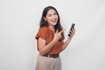 Happy Asian woman is smiling and pointing to her smartphone wearing brown shirt isolated by a white background.