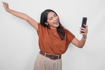 Fototapeta premium Joyful young Asian woman in brown shirt doing video call with happy successful expression and clenched fist gesture isolated over white background.