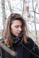 A young woman wearing a black coat and a black-and-white geometric scarf stands in front of a bare tree branches. She is looking away from the camera with a thoughtful expression on her face. - 794788047