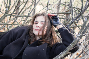 A young woman in a black coat is lying on a pile of branches in the forest. She has a pensive expression on her face and is looking off into the distance. The image is full of emotion and mystery. - 794788226