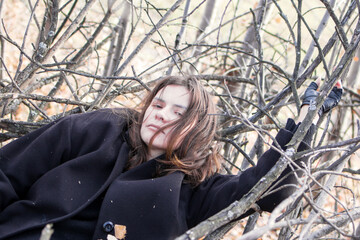 A young woman wearing a black coat and a black-and-white geometric scarf stands in front of a bare tree branches. She is looking away from the camera with a thoughtful expression on her face.