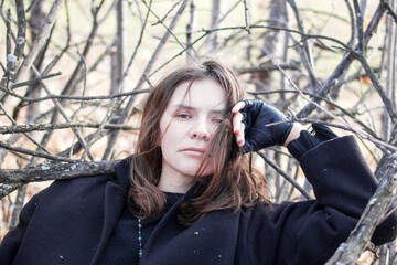 A young woman in a black coat is lying on a pile of branches in the forest. She has a pensive expression on her face and is looking off into the distance. The image is full of emotion and mystery. - 794788287