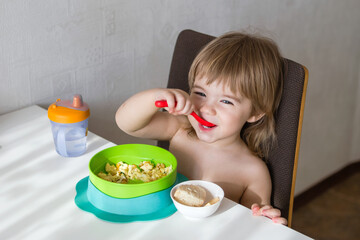 An adorable toddler happily sits at the table, enjoying breakfast. The childs cheerful face beams with joy as they hold a spoon and bowl, relishing each bite of cereal in a cozy kitchen setting.