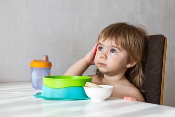 A young toddler girl with curly hair, dressed in pink overalls, sits at a wooden table. She looks thoughtfully at a plate, cup, and spoon set before her. Ideal for stock photo use. - 794788467