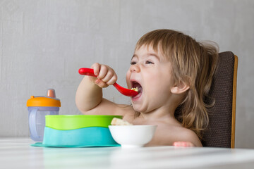 A cute little girl sits in a high chair and eats her breakfast. She is smiling and looks happy. The high chair is placed in a kitchen with a white wall in the background.