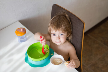A hungry toddler sits in a high chair and eats breakfast. The child holds a spoon and eats from a green bowl. There is bread on the table. Isolated on a white background.