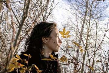 A young woman stands in a contemplative pose in the middle of an autumn forest. She is surrounded by colorful foliage. Her eyes closed and is wearing a black jacket.