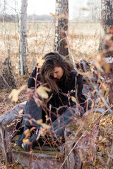 A young woman with long brown hair sits on the ground in a forest, eyes closed, smiling peacefully. She wears a black sweater and jeans. Tall grass and yellow leaves surround her. - 794788891