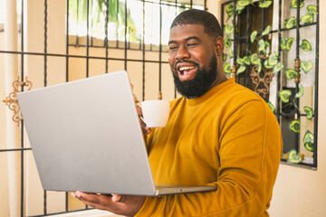 Black man relaxing at home in the morning with coffee enjoy using computer laptop addiction to technology trends following with emoji on social networks. Social medias and technology concept.