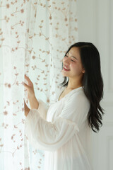 Asian woman in white pajamas is opening the curtains and looking out the window smiling in her room in the morning. Soft sunlight. Vacation, lifestyle concept.
