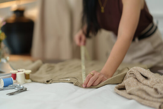 Confident Asian designer woman Beautiful smile using measuring tape for suit jacket with laptop computer in modern studio, clothes hanging on mannequin for customer online sales ordering.