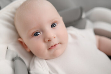  Cute infant lies in cocoon of a baby lounger, looks at camera, smiles, close-up. Portrait of blue-eyed newborn baby.Concept of maternity leave, child care, happy carefree childhood.