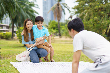 Happy cheerful Asian family with father, mother, and little son enjoy picnic together in a weekend at a park. Happy family moment concept. Parent and their child relax together in the park.