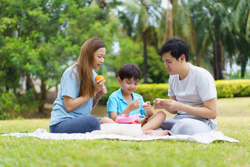 Naklejka na ściany i meble Happy cheerful Asian family with father, mother, and little son enjoy picnic together in a weekend at a park. Happy family moment concept. Parent and their child relax together in the park.