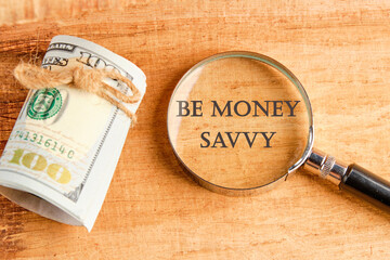 Savings concept,Text Be money savvy through a magnifying glass, they are visible on a beautiful background next to a roll of paper banknotes