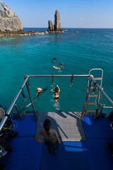 Tourists swimming in clear blue waters near a rocky island, seen from a boat deck