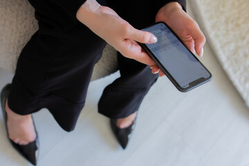 Woman holding mobile phone in hand and texting. Female hands and legs top view. Online chatting on smartphone of girl