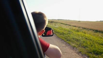 child boy son enjoys summer day with his hand out car window, car rushing along road, child feels...